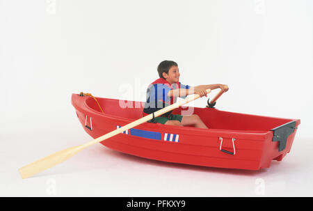 A boy wearing a life jacket sitting in a red rowing boat, holding paddles Stock Photo