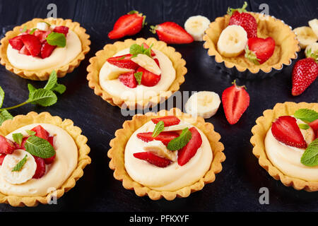 tartlets with strawberries, banana slices loaded with cream diplomat on a black slate tray, view from above, close-up Stock Photo