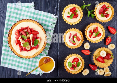 tartlets and tart with strawberries, banana slices loaded with custard cream on a black slate tray, with maple syrup in a bowl on wooden table, view f Stock Photo