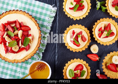 tartlets with strawberries, banana slices loaded with custard cream on a black slate plate. tart in a baking pan on textile mat view from above, close Stock Photo