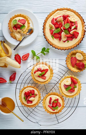 delicious ,tartlets with strawberries, banana slices loaded with custard cream on a metal grid and on a plate. tart in a baking pan white wooden table Stock Photo