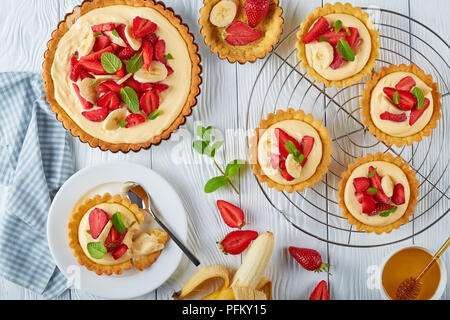 delicious tartlets with strawberries, banana slices loaded with custard cream on a metal grid and on a plate. tart in a baking pan white wooden table, Stock Photo