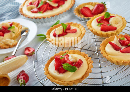 delicious tartlets with strawberries, banana slices loaded with custard cream on a metal grid and on a white plate with spoon, view from above, close- Stock Photo