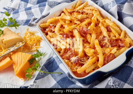 australian fries smothered in melted mix of shredded cheese and bacon in a baking dish on a white wooden table with ingredients at the background, vie Stock Photo