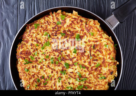delicious swiss rosti or potato pancake sprinkled with finely chopped parsley in a pan on a black wooden table, close-up, macro Stock Photo