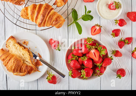 freshly baked croissants on a wire rack and a croissant has sliced on a plate to make a sandwich with fresh ripe strawberries and cream cheese, view f Stock Photo