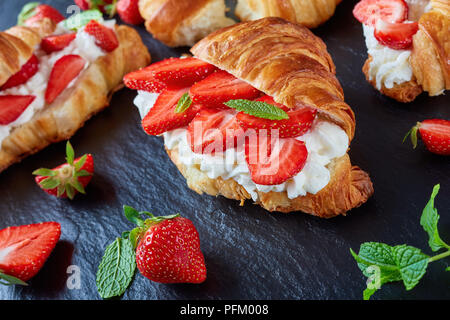 delicious french croissant sandwiches layered with fresh ripe strawberries and whipped cream cheese on a stone tray on a black wooden table, view from Stock Photo