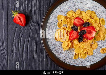 healthy breakfast - portion of chia seeds pudding topped with gluten free Corn flakes, fresh strawberry,and mulberries in a bowl on black wooden table Stock Photo
