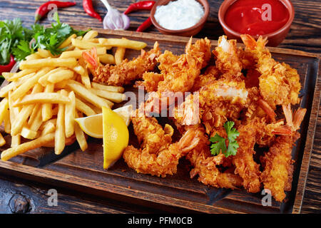 delicious crunchy corn flakes breaded and deep-fried shrimps with french fries on a cutting board, ketchup and cocktail sauce on a wooden table, view  Stock Photo