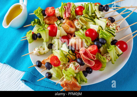 chicken meat shish kebab skewers with zucchini ribbons, tomatoes, mozzarella balls, salami slices, black olives on a white plate, horizontal view from Stock Photo