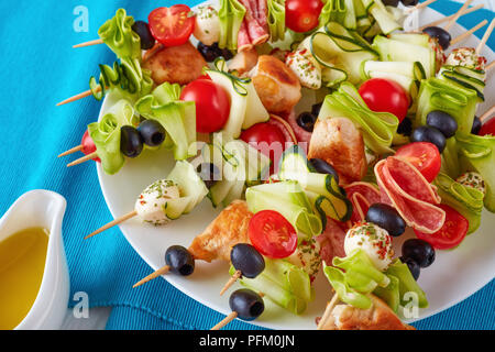 grilled chicken meat shish kebab skewers with zucchini ribbons, tomatoes, mozzarella balls, salami slices, black olives on a white plate, view from ab Stock Photo