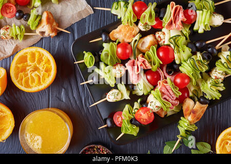 antipasto skewers with chicken meat, zucchini, cherry tomatoes,  mozzarella balls, salami slices, olives on a black plate on a wooden table with orang Stock Photo