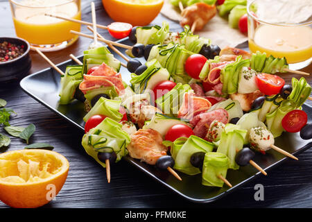 kebab on skewers with chicken meat, zucchini, tomatoes, mozzarella balls, salami slices, olives on a black plate on a wooden table with fresh orange j Stock Photo