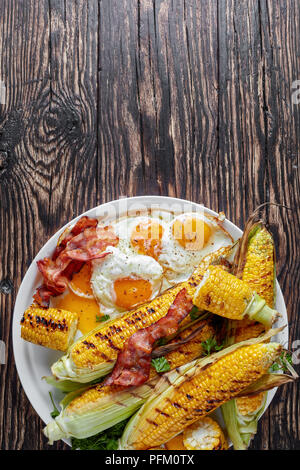 tasty grilled corn in cobs with fried eggs and bacon slices on a white dish on an old dark rustic table, vertical view from above, flat lay Stock Photo