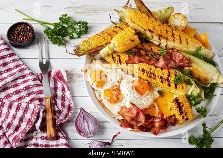 grilled corn in cobs with fried eggs and bacon slices on a white dish, on an old rustic table, view from above, close-up Stock Photo
