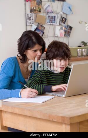 Boy using laptop, woman sitting next to him with pen and notebook Stock Photo