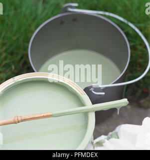 Pot of pale green paint, wooden stirring stick resting across top, bucket of green paint behind, on green grass. Stock Photo