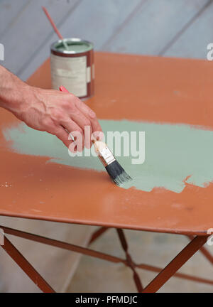 Hand painting wooden garden table with pale green paint, using paintbrush, paint pot with stirring stick in background. Stock Photo