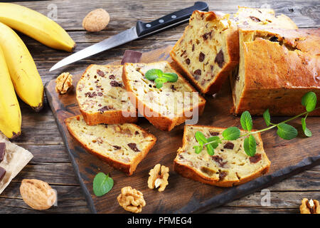 freshly baked delicious banana bread with walnuts and chocolate pieces cut in slices on cutting board. ingredients on rustic wooden table, american cu Stock Photo