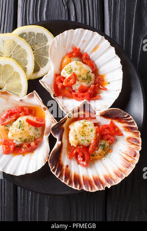 sea scallops in a shell with sauce, tomatoes, parsley and lemon close-up on a dark background. vertical top view from above Stock Photo