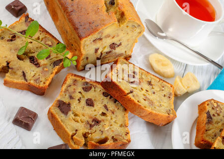 close-up of freshly baked delicious banana bread with walnuts and chocolate pieces cut in slices on cutting board. ingredients and cup of tea on woode Stock Photo