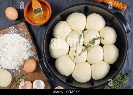 unbaked soft and fluffy dinner buns in baking dish with dough, flour and egg on a cutting board, olive oil, rolling pin on wooden kitchen table, view  Stock Photo