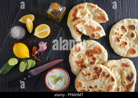 pan fried homemade hot pita bread on black wooden table,  fresh tzatziki sauce in bowl and ingredients on stone cutting board, view from above Stock Photo