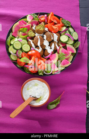delicious falafel balls on plate with vegetable colorful salad of watermelon radish, chard leaves, cucumber and tomato slices. tzatziki sauce in bowl, Stock Photo