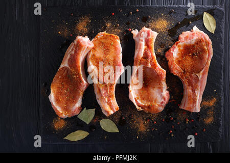 raw pork chops seasoned with spices, peppercorns and bay leaf on a black slate tray on wooden table, view from above Stock Photo