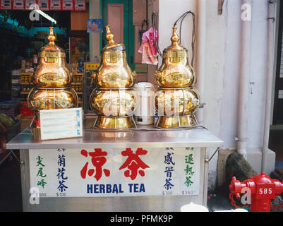 Singapore, Chinatown, Temple Street, decorative herbal tea dispensers, gold coloured metal decorated with Chinese script and dragons, lid loosely placed on central dispenser, standing on metal box, shop in background. Stock Photo