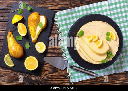 homemade pear sponge cake with cream cheese, decorated with mint, pear and lemon slices on black slate plate on napkin with spatula and ingredients at Stock Photo