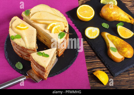 delicious sliced homemade pear sponge cake with cream cheese, decorated with mint, pear slices and lemon on black slate plate. ingredients on cutting  Stock Photo