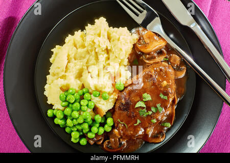 portion of hot savory juicy salisbury steaks with mushroom onion gravy served with mashed potatoes and green peas on black plates with cutlery, restau Stock Photo