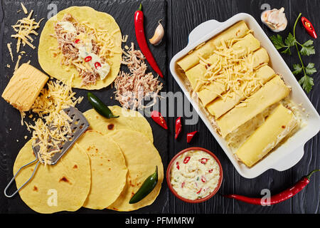 cooking enchiladas - homemade corn tortillas layered with shredded meat, sauce, grated cheese and spices in baking dish and on black stone board,  vie Stock Photo