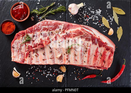 close-up of rack of uncooked pork spare ribs seasoned with spices on black slate tray with paprika, garlic cloves, bay leaves at background, view from Stock Photo