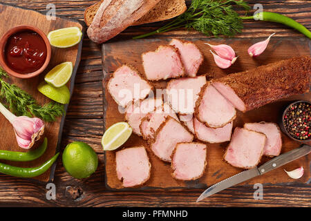 delicious sliced ham seasoned with ground spices served on wooden cutting board with barbecue sauce, lime slices, garlic and rye bread at background,  Stock Photo