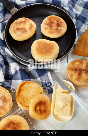 hot english muffins on a dry skillet and on a grid on wooden table with kitchen towel, fresh butter and knife, view from above, close-up Stock Photo