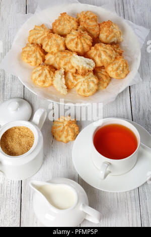 delicious homemade coconut cookies on a white plate on wooden table with cup of tea, sugar bowl and fresh cream in a milk jug at background, vertical  Stock Photo