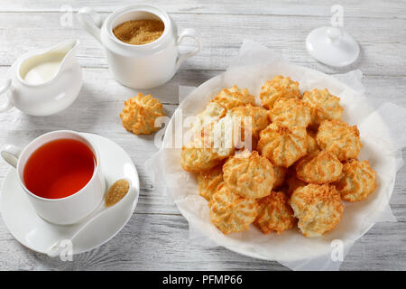 delicious homemade coconut cookies on a white plate on wooden table with cup of tea, sugar bowl and fresh cream in a milk jug at background, horizonta Stock Photo