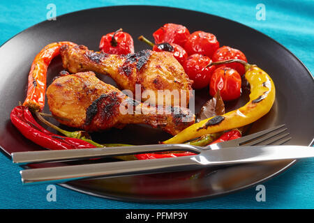 portion of delicious grilled chicken drumsticks with chili peppers and cherry tomatoes served on a black plate with silver fork and knife, horizontal  Stock Photo
