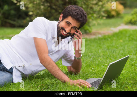 Young happy smiling man working with laptop and cellphone, outdoors Stock Photo