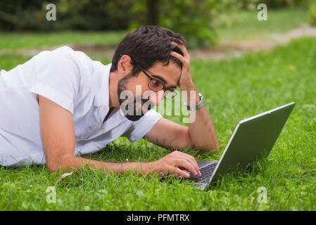 Young man working on his laptop while lying on the grass of the park. Happy expression, concept of freedom. Stock Photo