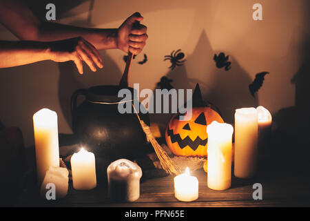 witch hand boiling potion for spell, with jack o lantern pumpkin with candles, broom and bats, ghosts on background in dark spooky room. Happy Hallowe Stock Photo