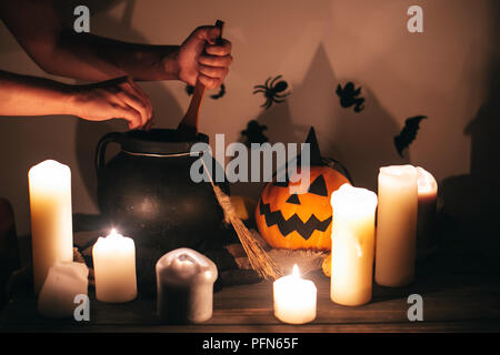 witch hand boiling potion for spell, with jack o lantern pumpkin with candles, broom and bats, ghosts on background in dark spooky room. Happy Hallowe Stock Photo