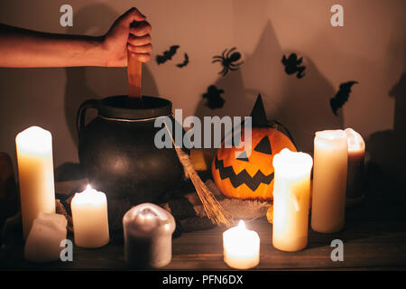 witch hand boiling potion for spell, with jack o lantern pumpkin with candles, broom and bats, ghosts on background in dark spooky room. Happy Hallowe Stock Photo