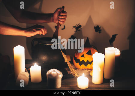 witch hand boiling potion for spell, with jack o lantern pumpkin with candles, broom and bats, ghosts on background in dark spooky room. Happy Hallowe Stock Photo