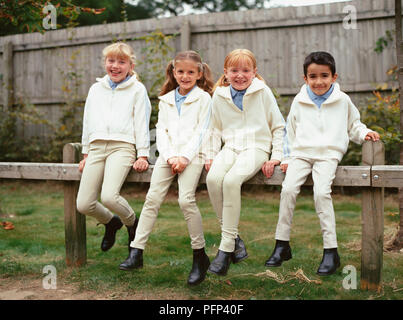 Three young girls and a boy sitting on a fence, wearing horse riding clothes. Stock Photo