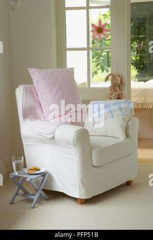 Armchair with two cushions on it, next to small side table with glass of water and biscuits on it, soft toy on window sill Stock Photo