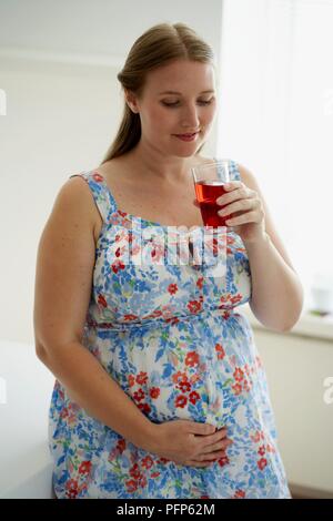 Smiling pregant woman standing with hand on belly, holding glass of fruit juice in left hand Stock Photo