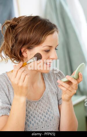 Young woman applying blusher to her face while holding pocket mirror in the other hand Stock Photo